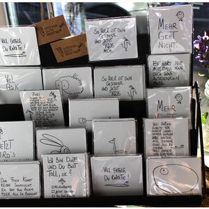 Puede incluir: Exhibici&oacute;n de tarjetas blancas con texto manuscrito negro e ilustraciones sencillas. Las tarjetas est&aacute;n en fundas de pl&aacute;stico y est&aacute;n dispuestas en un estante de metal negro. Algunas tarjetas tienen frases como "Vel Erfolg, Du Rakete!" y "Mehr Geht Nicht."