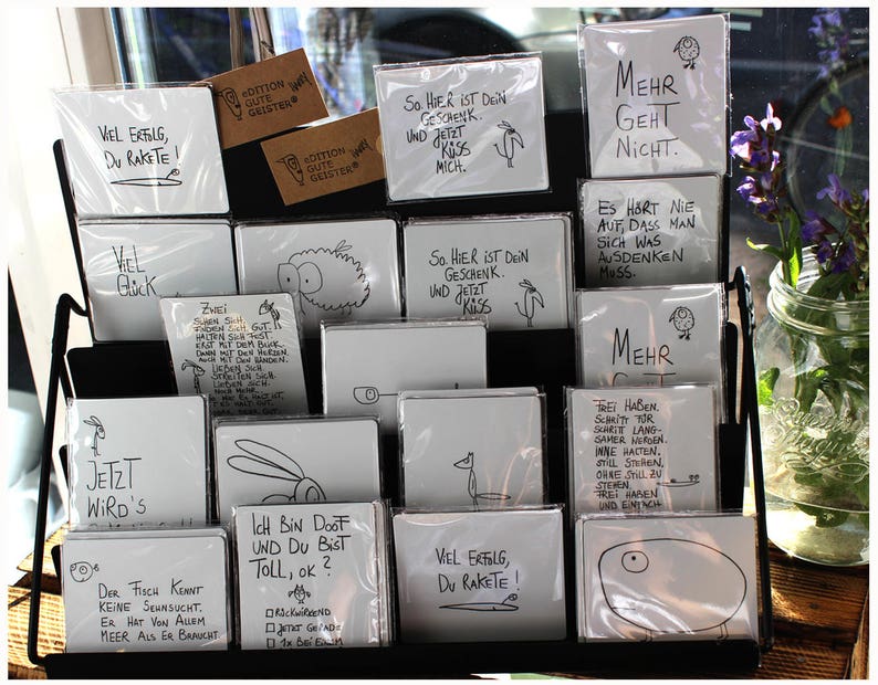 Puede incluir: Exhibici&oacute;n de tarjetas blancas con texto manuscrito negro e ilustraciones sencillas. Las tarjetas est&aacute;n en fundas de pl&aacute;stico y est&aacute;n dispuestas en un estante de metal negro. Algunas tarjetas tienen frases como "Vel Erfolg, Du Rakete!" y "Mehr Geht Nicht."