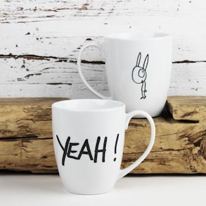 Könnte beinhalten: Zwei weiße Keramikbecher. Ein Becher hat das Wort "YEAH!" in schwarzer Schrift. Der andere Becher zeigt eine skurrile schwarze Strichzeichnung eines Kaninchens. Die Becher stehen auf einer Holzoberfläche vor einem weißen Hintergrund.