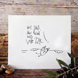 Könnte beinhalten: Eine quadratische weiße Grußkarte mit einer schwarzen Strichzeichnung einer fliegenden Eule und dem handschriftlichen deutschen Text "Viel Spaß, hau rein und volle Pulle!" mit Ausrufezeichen, platziert auf einer rustikalen Holzoberfläche mit Steinen.