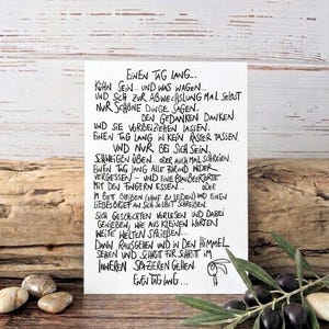 Könnte beinhalten: Eine handgeschriebene Notiz mit dem Text "Einen Tag Lang..." in schwarzer Tinte. Die Notiz befindet sich auf weißem Papier mit einer kleinen Kaninchenillustration. Der Hintergrund ist eine Holzoberfläche mit Oliven und Steinen.