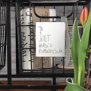 Puede incluir: Unidad de almacenamiento de metal negro con cestas de alambre que contiene papel enrollado y un letrero que dice "JETZT WIRD'S ROMANTISCH." Dos tulipanes naranjas y rosas est&aacute;n en primer plano.