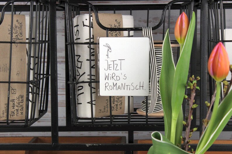 Puede incluir: Unidad de almacenamiento de metal negro con cestas de alambre que contiene papel enrollado y un letrero que dice "JETZT WIRD'S ROMANTISCH." Dos tulipanes naranjas y rosas est&aacute;n en primer plano.