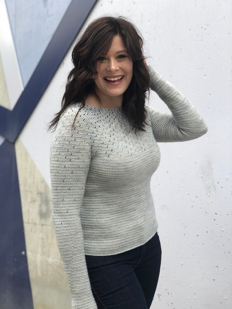 May include: A light gray sweater with a blue speckled pattern. The sweater has long sleeves and a round neckline.