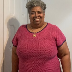 May include: A woman wearing a pink crocheted top with a blue trim and a gold necklace with a sun pendant.