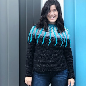 Könnte beinhalten: Eine Frau trägt einen schwarzen und blauen Strickpullover mit einem einzigartigen Design. Der Pullover hat einen hohen Kragen und lange Ärmel.