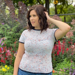 May include: A white crochet top with a speckled pattern in red, blue, and white. The top has short sleeves and a navy blue trim around the neckline and sleeves.
