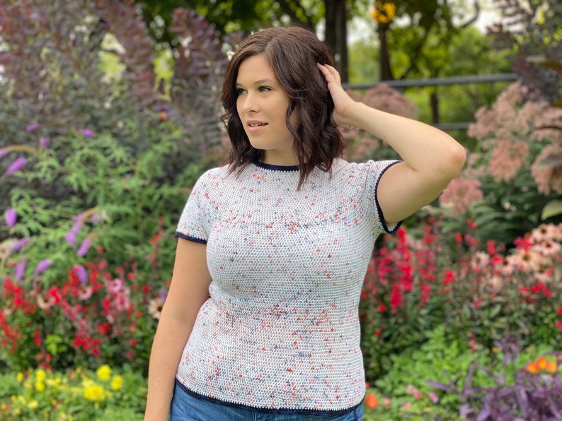 May include: A white crochet top with a speckled pattern in red, blue, and white. The top has short sleeves and a navy blue trim around the neckline and sleeves.