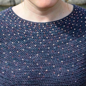 May include: A close-up of a blue crocheted sweater with a beaded neckline. The beads are a mix of white and brown.