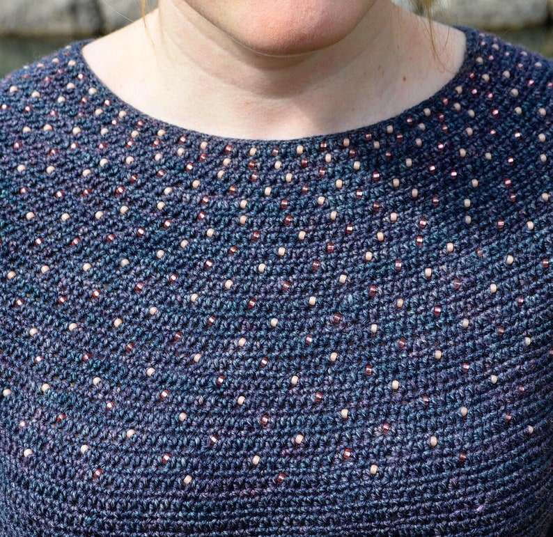May include: A close-up of a blue crocheted sweater with a beaded neckline. The beads are a mix of white and brown.