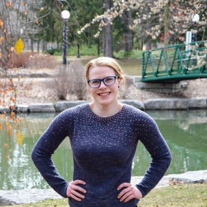 May include: A woman with short, blonde hair is wearing a blue sweater with a subtle sparkle pattern. She is smiling and looking at the camera. The sweater has a round neckline and long sleeves.