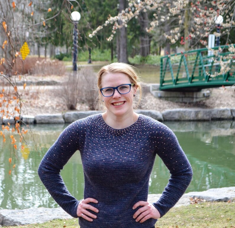May include: A woman with short, blonde hair is wearing a blue sweater with a subtle sparkle pattern. She is smiling and looking at the camera. The sweater has a round neckline and long sleeves.