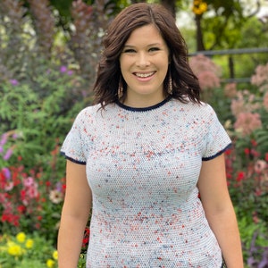 May include: A woman with brown hair is smiling and wearing a white, blue, and red speckled short-sleeved shirt with a navy blue trim.