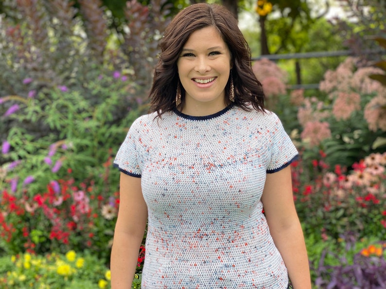 May include: A woman with brown hair is smiling and wearing a white, blue, and red speckled short-sleeved shirt with a navy blue trim.