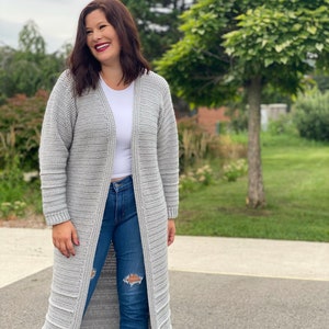 May include: A long, gray cardigan with a ribbed texture. The cardigan is open in the front and has long sleeves. The person wearing the cardigan is smiling and looking to the right.