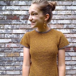 May include: A woman wearing a mustard yellow crochet top with short sleeves and a grey trim. The top has a textured, open-stitch design.