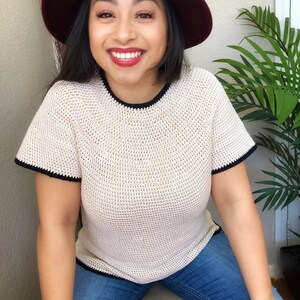 May include: A woman wearing a white crocheted top with black trim, blue jeans, and a burgundy fedora.