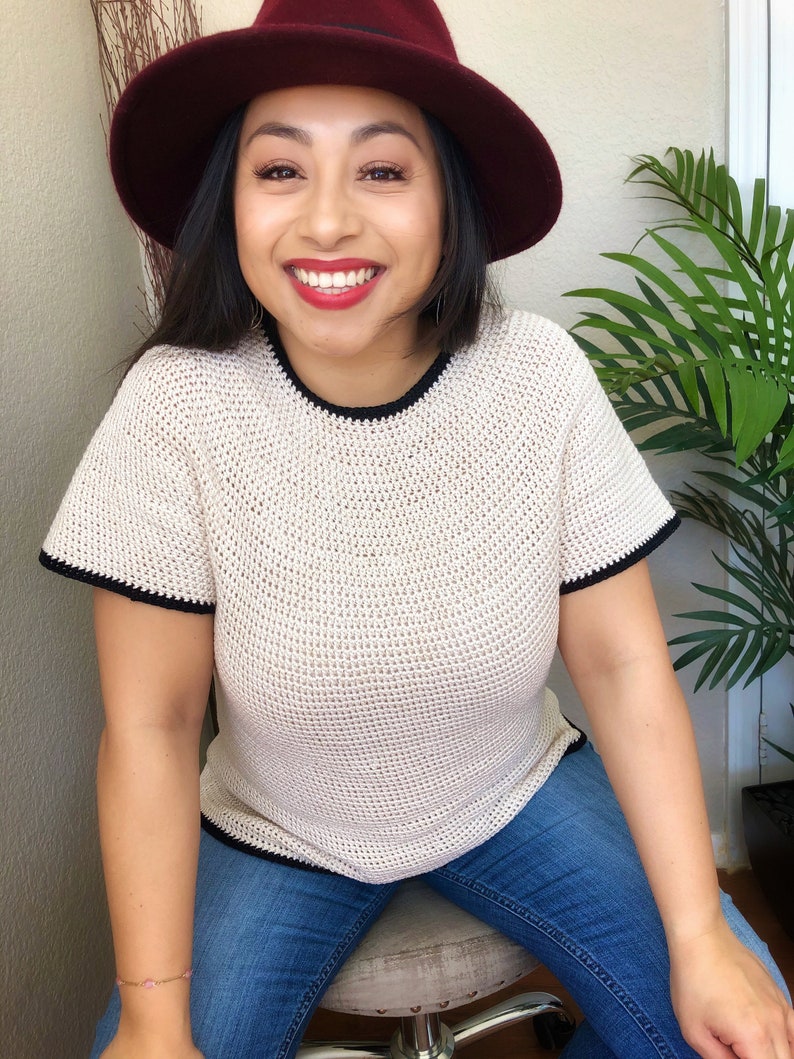 May include: A woman wearing a white crocheted top with black trim, blue jeans, and a burgundy fedora.