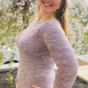 May include: A close-up of a woman wearing a light purple crocheted sweater with a textured pattern. The sweater has long sleeves and a round neckline.