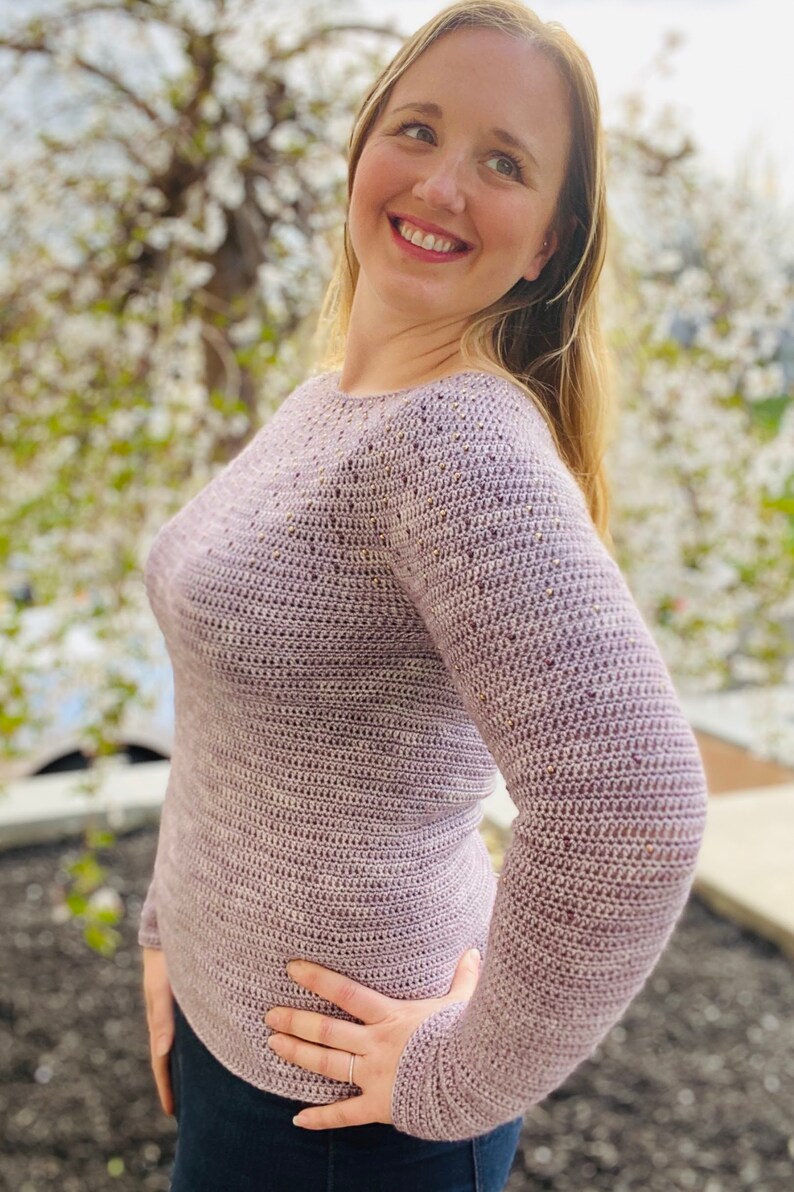 May include: A close-up of a woman wearing a light purple crocheted sweater with a textured pattern. The sweater has long sleeves and a round neckline.