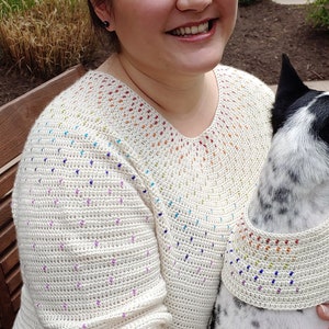 May include: A woman wearing a white crocheted sweater with a rainbow design and blue jeans. She is holding a black and white dog wearing a matching crocheted collar.