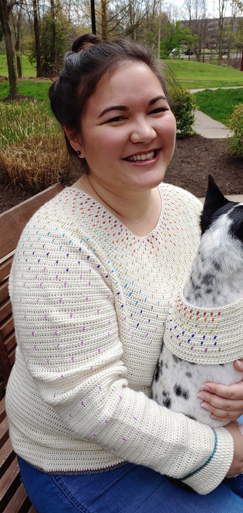 May include: A woman wearing a white crocheted sweater with a rainbow design and blue jeans. She is holding a black and white dog wearing a matching crocheted collar.