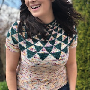 May include: A woman wearing a short-sleeved, multi-colored crochet top with a geometric pattern in shades of green, purple, and white. The top has a high neckline and a fitted silhouette.