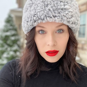May include: A woman wearing a grey faux fur headband and a black turtleneck sweater. She has long dark hair and is looking directly at the camera. Her lips are painted a bright red.