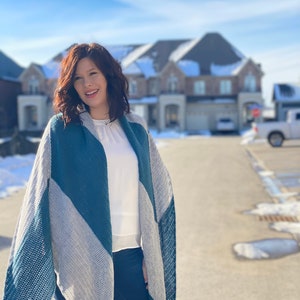 May include: A woman wearing a blue and grey striped knitted scarf, a white long-sleeved shirt, and blue jeans. She is standing in front of a row of houses.