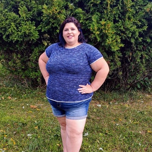 May include: A woman wearing a blue and white knit top with short sleeves and denim shorts. She is standing in front of a green bush.