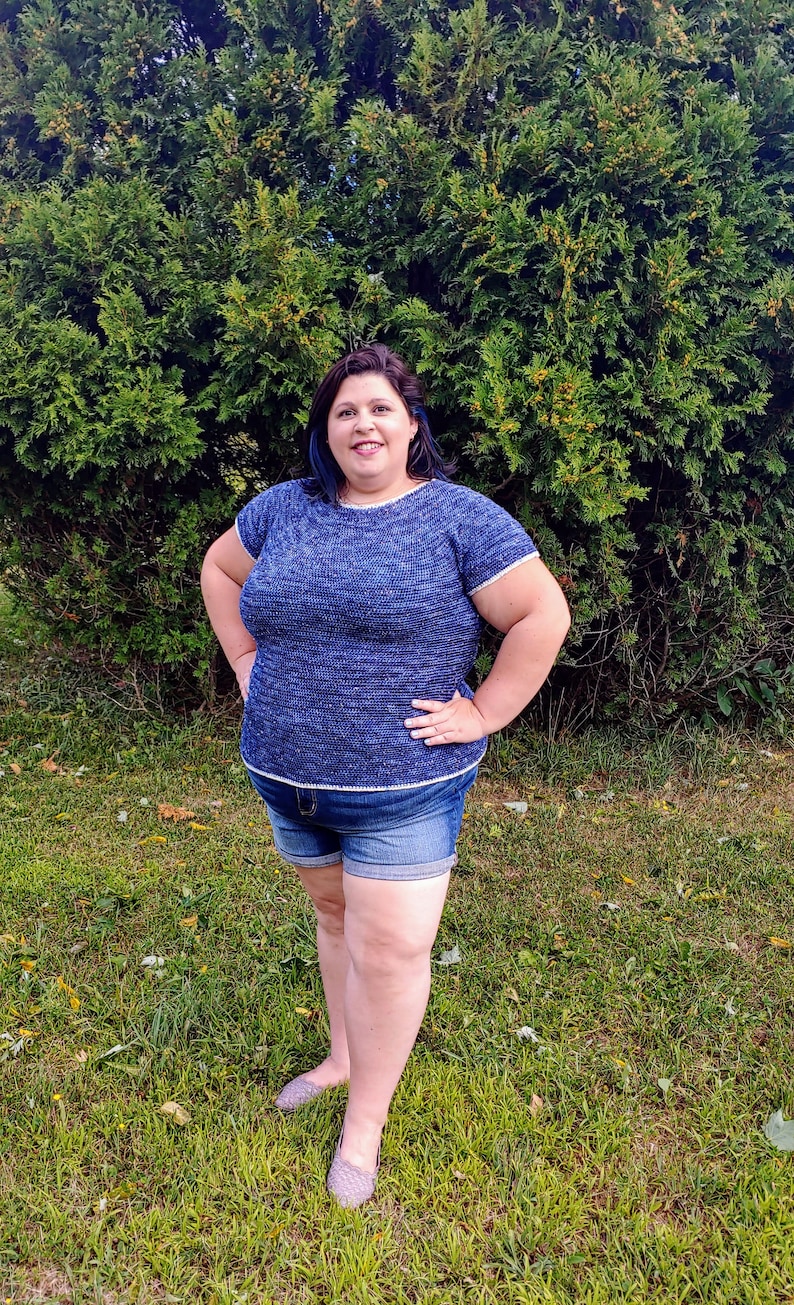 May include: A woman wearing a blue and white knit top with short sleeves and denim shorts. She is standing in front of a green bush.