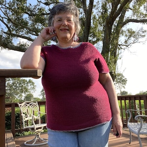 May include: A woman wearing a maroon crocheted top with a blue and white trim, blue jeans, and glasses. She is standing on a wooden deck with a tree in the background.