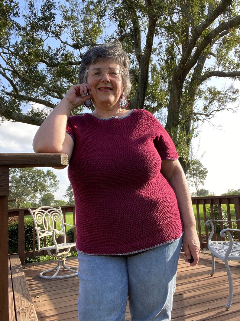 May include: A woman wearing a maroon crocheted top with a blue and white trim, blue jeans, and glasses. She is standing on a wooden deck with a tree in the background.