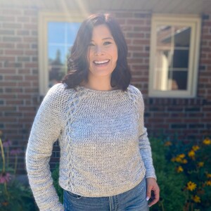 May include: A woman wearing a light gray crocheted sweater with a braided detail down the front. She is smiling and looking at the camera. The sweater has long sleeves and a round neckline.