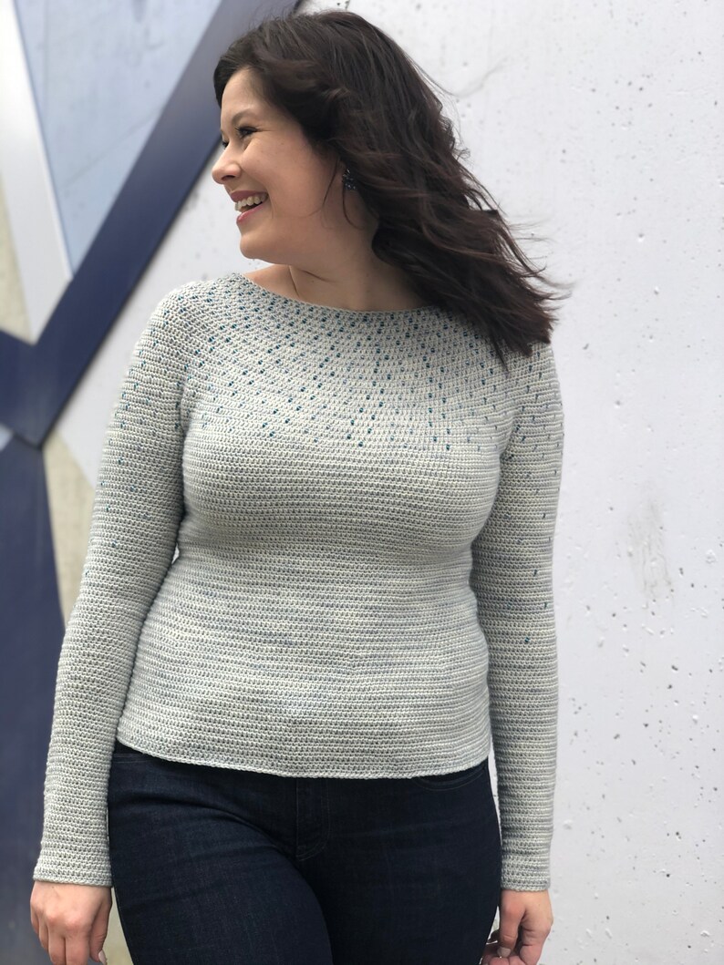 May include: A light gray crochet sweater with a blue speckled pattern. The sweater has long sleeves and a round neckline.