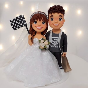 May include: A wedding cake topper featuring a bride and groom. The bride is wearing a white wedding dress and holding a checkered flag. The groom is wearing a grey jacket, a black shirt with the word "VIPER" on it, and holding a brown paper bag.