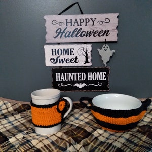 Puede incluir: Una taza y un tazón blancos con cubiertas de punto de crochet a rayas naranja y negras. La taza y el tazón están sobre una manta a cuadros. Un letrero con el texto "Happy Halloween", "Home Sweet" y "Haunted Home" está colgado en una pared en el fondo.