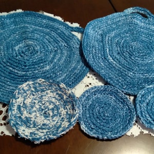Puede incluir: Cuatro posavasos redondos de ganchillo azul y blanco. Dos son más grandes y tienen asas, dos son más pequeños. Están sobre un mantel de encaje blanco.