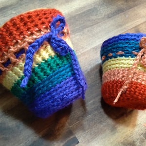 May include: Two crocheted rainbow-colored plant pots with purple, blue, green, yellow, pink, and red yarn. The larger pot has a purple ribbon tied around it, and the smaller pot has a pink ribbon tied around it.