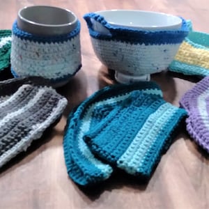 Puede incluir: Una colección de artículos de ganchillo hechos a mano en varios colores, incluyendo verde, gris, azul, amarillo y morado. Los artículos incluyen fundas para tazas, fundas para cuencos y agarraderas, todos con diseños a rayas. Dispuestos sobre una superficie de madera.