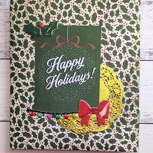 May include: A festive holiday card with a green background featuring a holly pattern. The card includes a green rectangle with the text "Happy Holidays!" and is decorated with a red bow, a gold doily, and colorful lights.