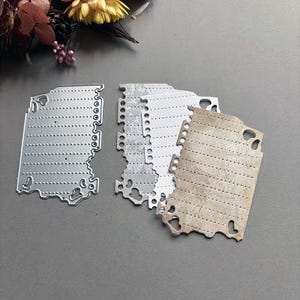 Puede incluir: Un conjunto de troqueles metálicos con forma de página de bloc de notas. Los troqueles son plateados y beige, con líneas punteadas y bordes decorativos. Estos suministros de manualidades son ideales para proyectos de scrapbooking y manualidades en papel.