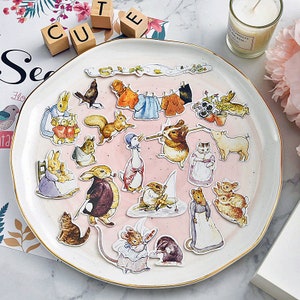 May include: A white plate with a pink background features a collection of colorful stickers featuring characters from the children's book, "The Tale of Peter Rabbit". The stickers include Peter Rabbit, Benjamin Bunny, Mrs. Tiggy-Winkle, and others.