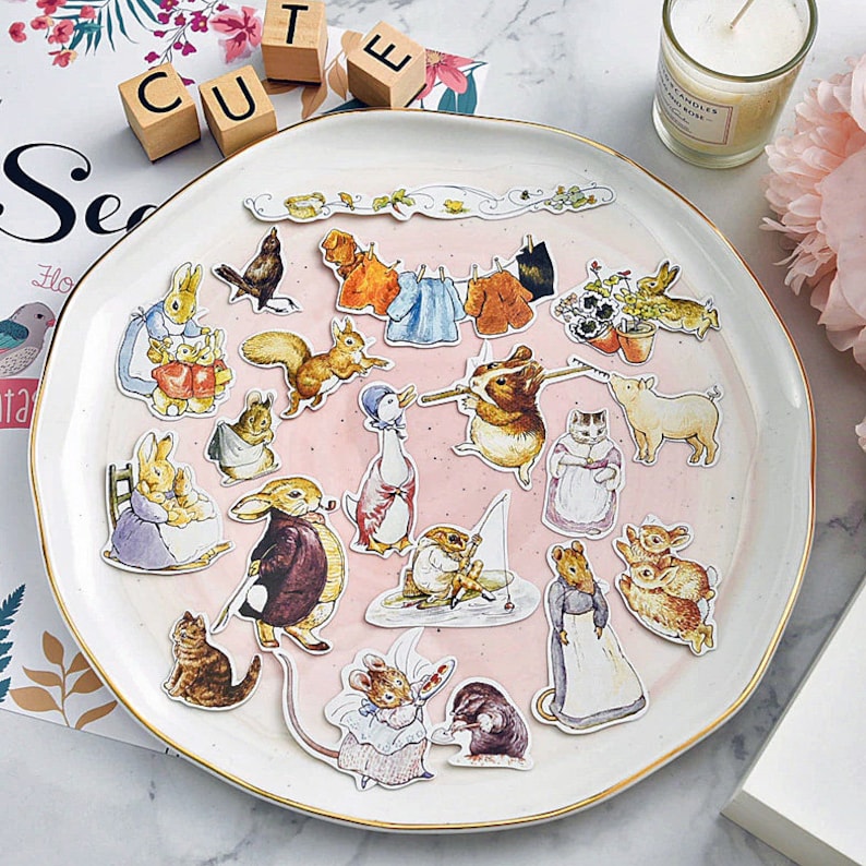 May include: A white plate with a pink background features a collection of colorful stickers featuring characters from the children's book, "The Tale of Peter Rabbit". The stickers include Peter Rabbit, Benjamin Bunny, Mrs. Tiggy-Winkle, and others.