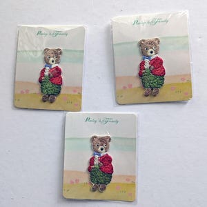 Op de afbeelding: Drie geborduurde strijkpatches met een bruine beer die een rood jasje en een groene broek draagt. Elke patch is op een witte kaart met de tekst "Pawley's Family" erop gedrukt.