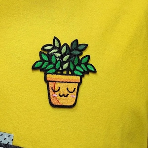 May include: An embroidered patch featuring a potted plant with a cute cartoon face. The plant has green leaves and the pot is yellow with a brown rim.