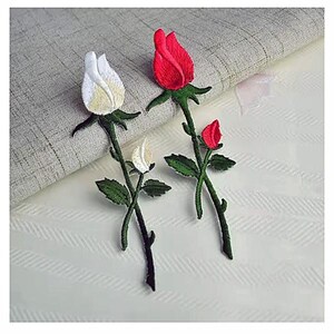 Può includere: Toppe di rose ricamate in rosso e bianco. Le toppe presentano steli e foglie verdi, con un design a punto dettagliato. Le rose sono in varie fasi di fioritura, con una completamente aperta e altre in bocciolo. Le toppe sono su uno sfondo chiaro.