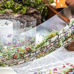 May include: Transparent washi tape with a detailed illustration of a stone wall and vibrant wildflowers. The tape's design is visible, with a repeating floral pattern. The tape is ideal for crafts and scrapbooking. The tape roll is visible.