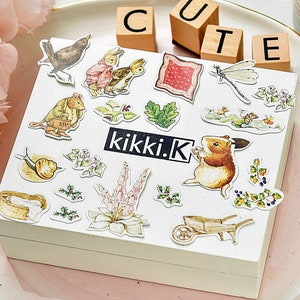 May include: A sheet of colorful stickers featuring various illustrations, including a snail, a mouse, a rabbit, a dragonfly, a flower, a wheelbarrow, and a fox. The stickers are on a white background with the text "kikki.K" printed in black.