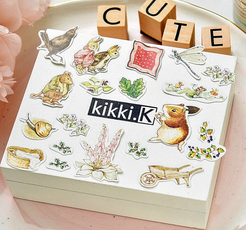 May include: A sheet of colorful stickers featuring various illustrations, including a snail, a mouse, a rabbit, a dragonfly, a flower, a wheelbarrow, and a fox. The stickers are on a white background with the text "kikki.K" printed in black.